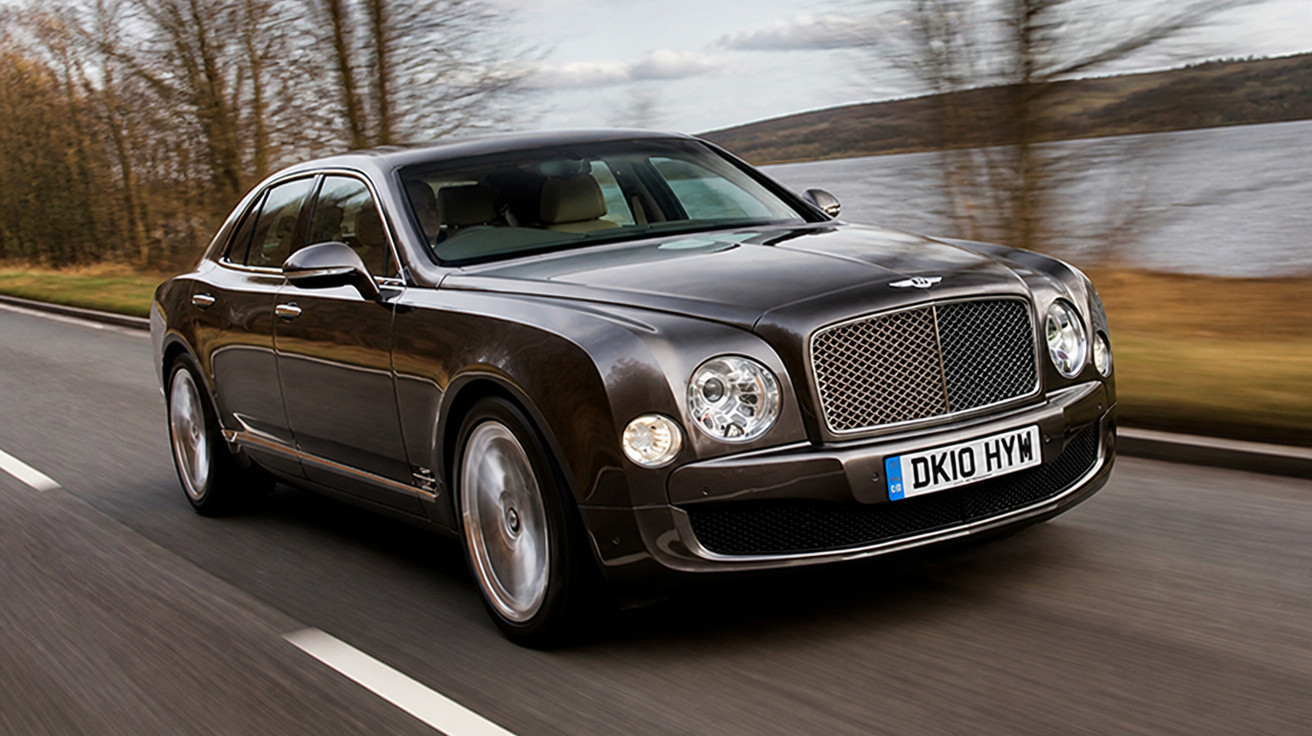 Carro Bentley preto luxo a circular numa estrada perto de um rio e árvores sem folhas.