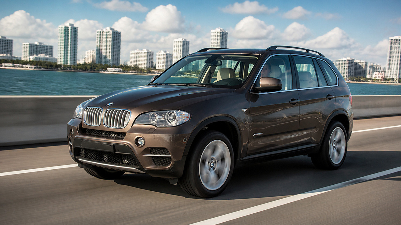 SUV BMW X5 castanho a circular numa estrada junto ao mar com arranha-céus ao fundo e céu azul com nuvens.