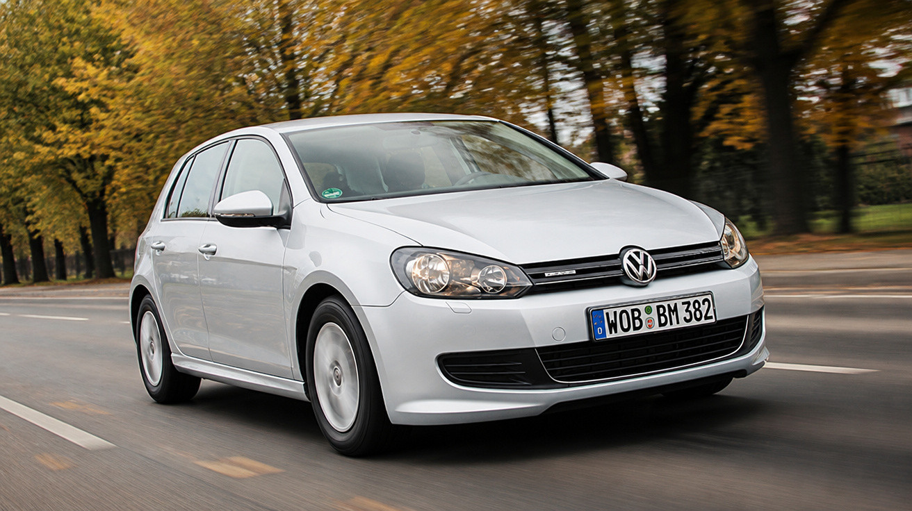 Carro Volkswagen Golf branco a conduzir numa estrada com árvores de folhas amarelas ao fundo.