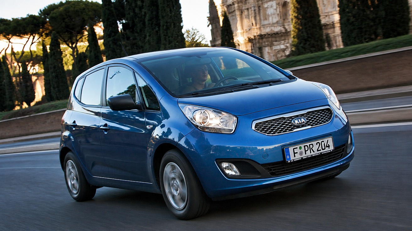 Carro azul Kia em movimento numa estrada com árvores e edifício antigo ao fundo.