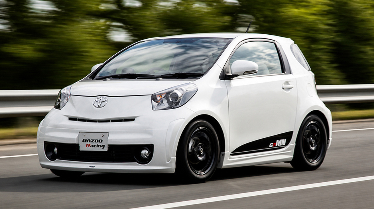 Carro compacto branco Toyota Gazoo Racing em movimento numa estrada com barreira metálica e vegetação.