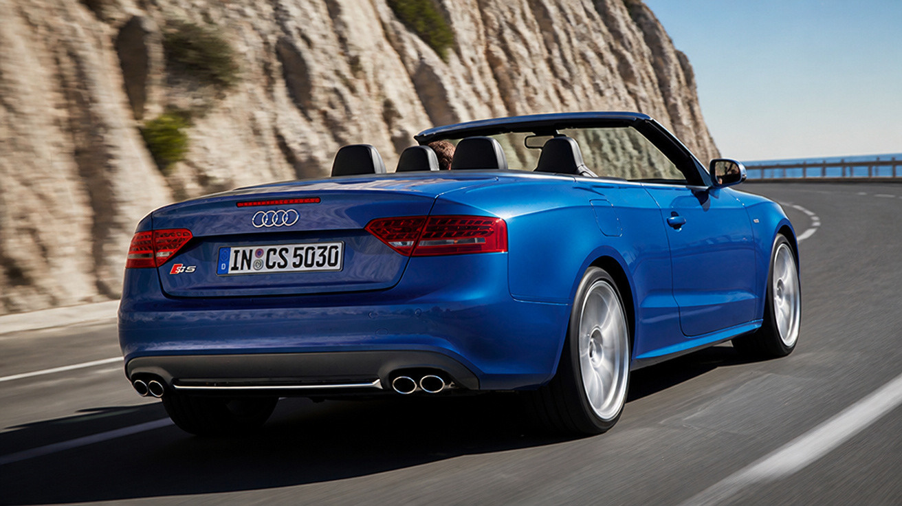 Automóvel descapotável Audi azul em estrada sinuosa junto a falésia rochosa sob céu limpo.
