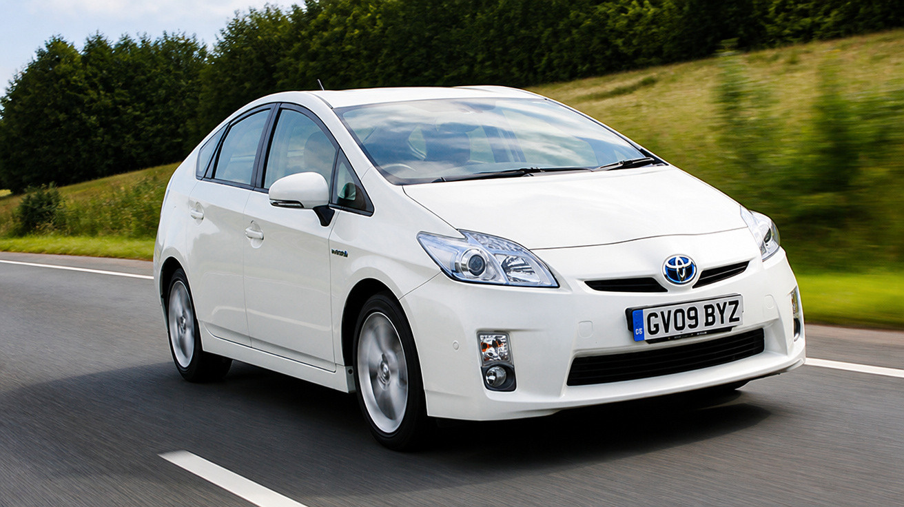 Carro híbrido branco Toyota Prius em movimento numa estrada asfaltada com vegetação ao fundo.