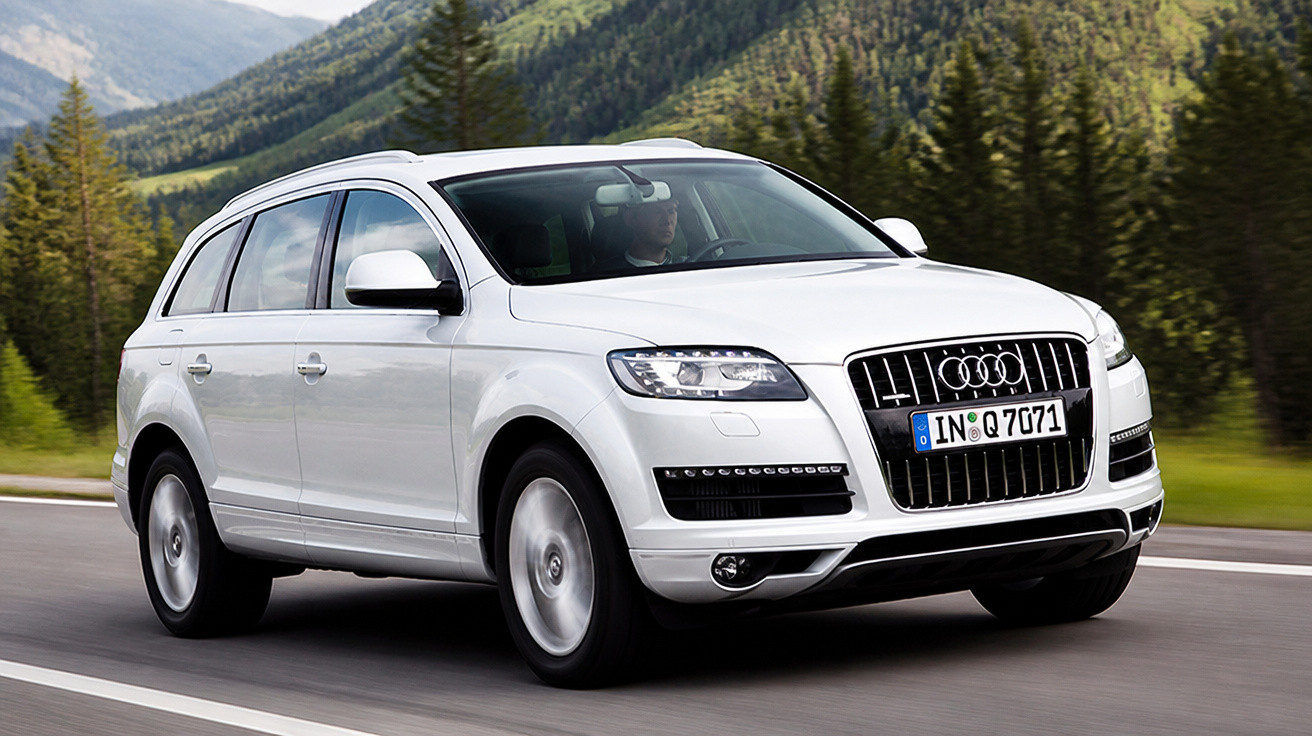 Carro Audi Q7 branco a circular numa estrada rodeada por montanhas e árvores verdes.