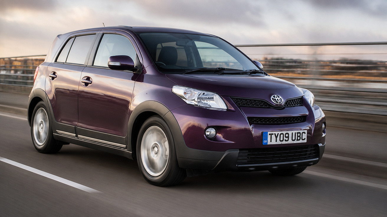 Carro Toyota Roxo a circular numa estrada com vedação metálica em ambiente urbano ao pôr do sol.