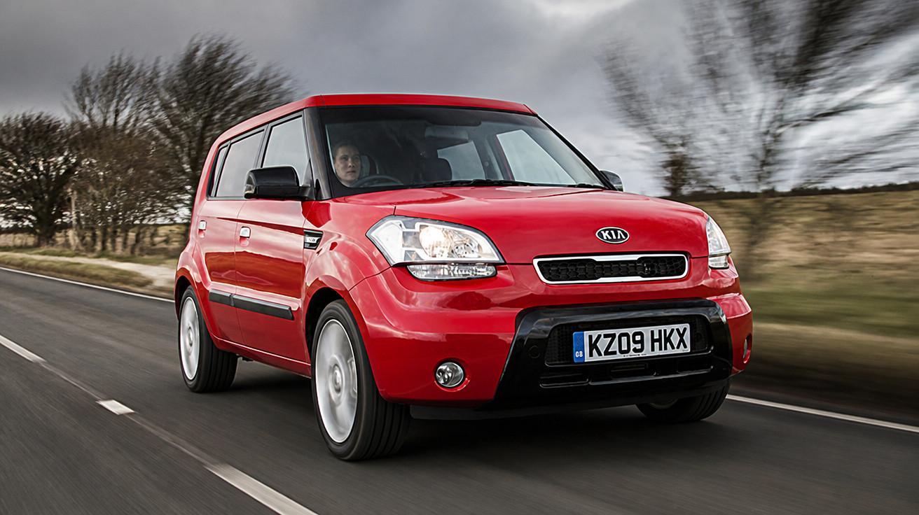 Carro vermelho Kia Soul a circular numa estrada rural com árvores e céu nublado ao fundo.