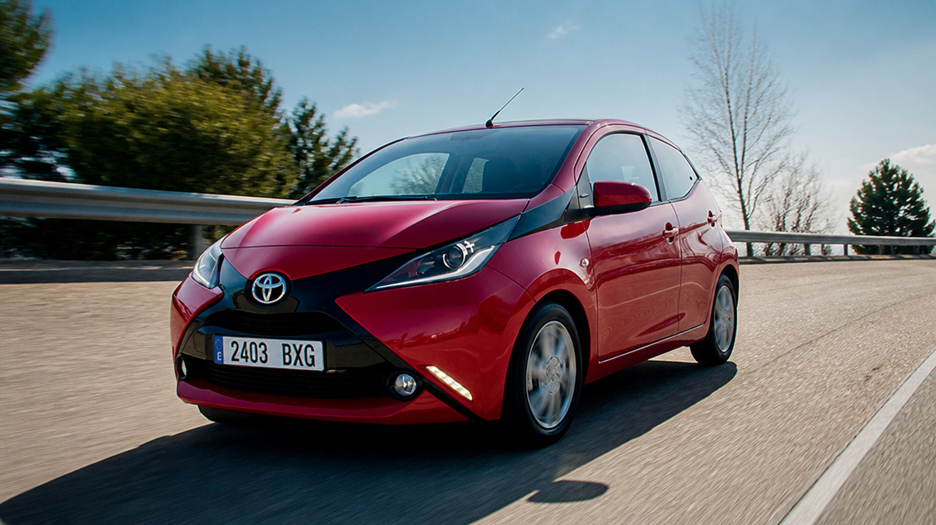 Carro Toyota Aygo vermelho a circular numa estrada sob céu azul e árvores ao fundo.