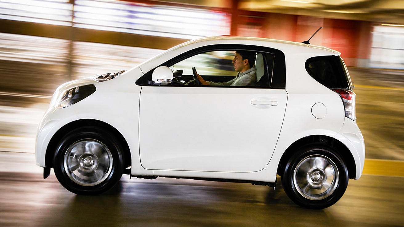 Carro branco pequeno em movimento com condutor masculino num ambiente urbano ao anoitecer.