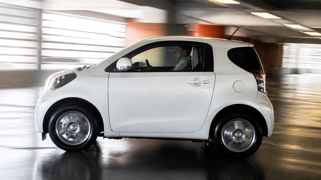 Carro branco compacto em movimento dentro de um parque de estacionamento coberto com um condutor visível.
