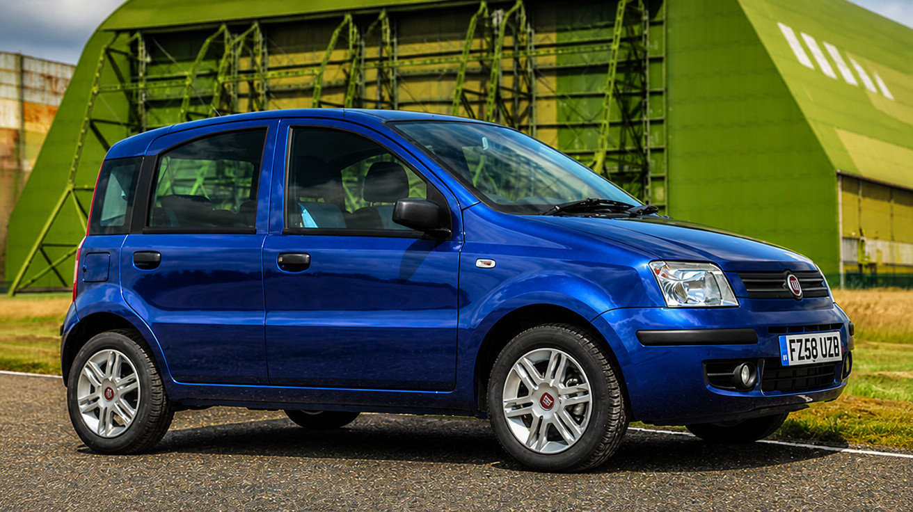 Carro compacto azul estacionado em estrada com armazém verde ao fundo.