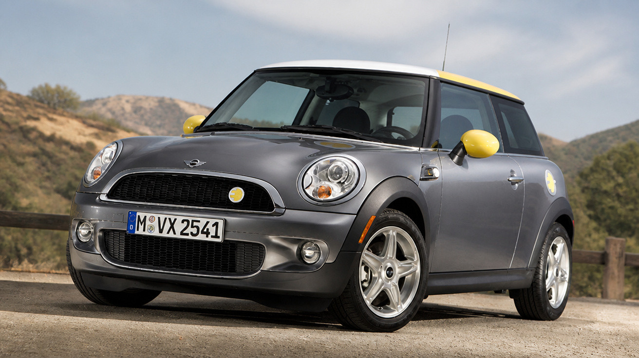 Carro Mini Cooper cinzento com detalhes amarelos estacionado numa estrada com montanhas ao fundo.