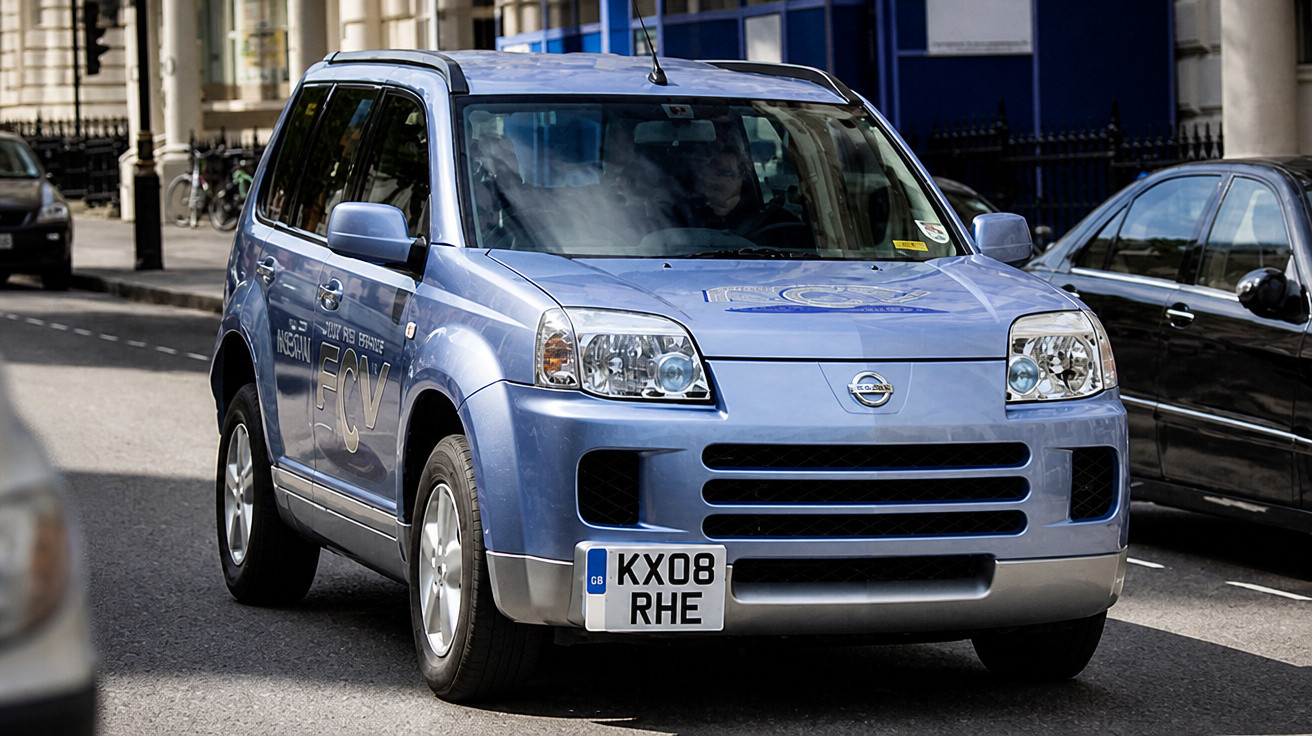 Carro azul Nissan X-Trail FCV estacionado numa rua urbana com veículos ao redor.