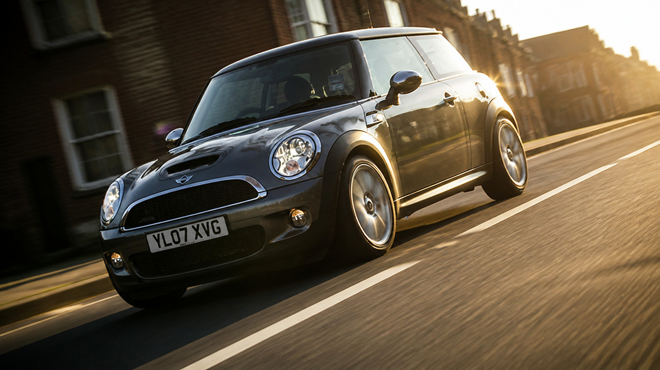 Carro Mini Cooper cinza escuro a circular numa estrada urbana ao pôr do sol.