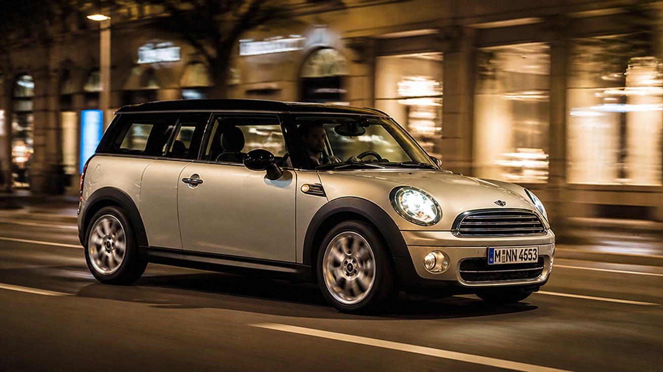 Carro Mini Cooper cinzento em movimento à noite numa rua urbana iluminada.