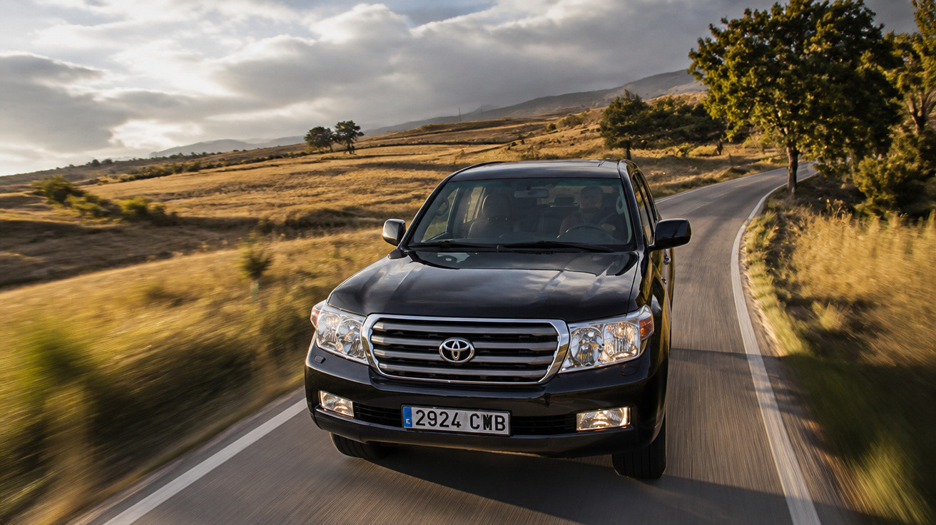 Carro Toyota Land Cruiser preto a circular numa estrada rural entre campos e árvores ao entardecer.