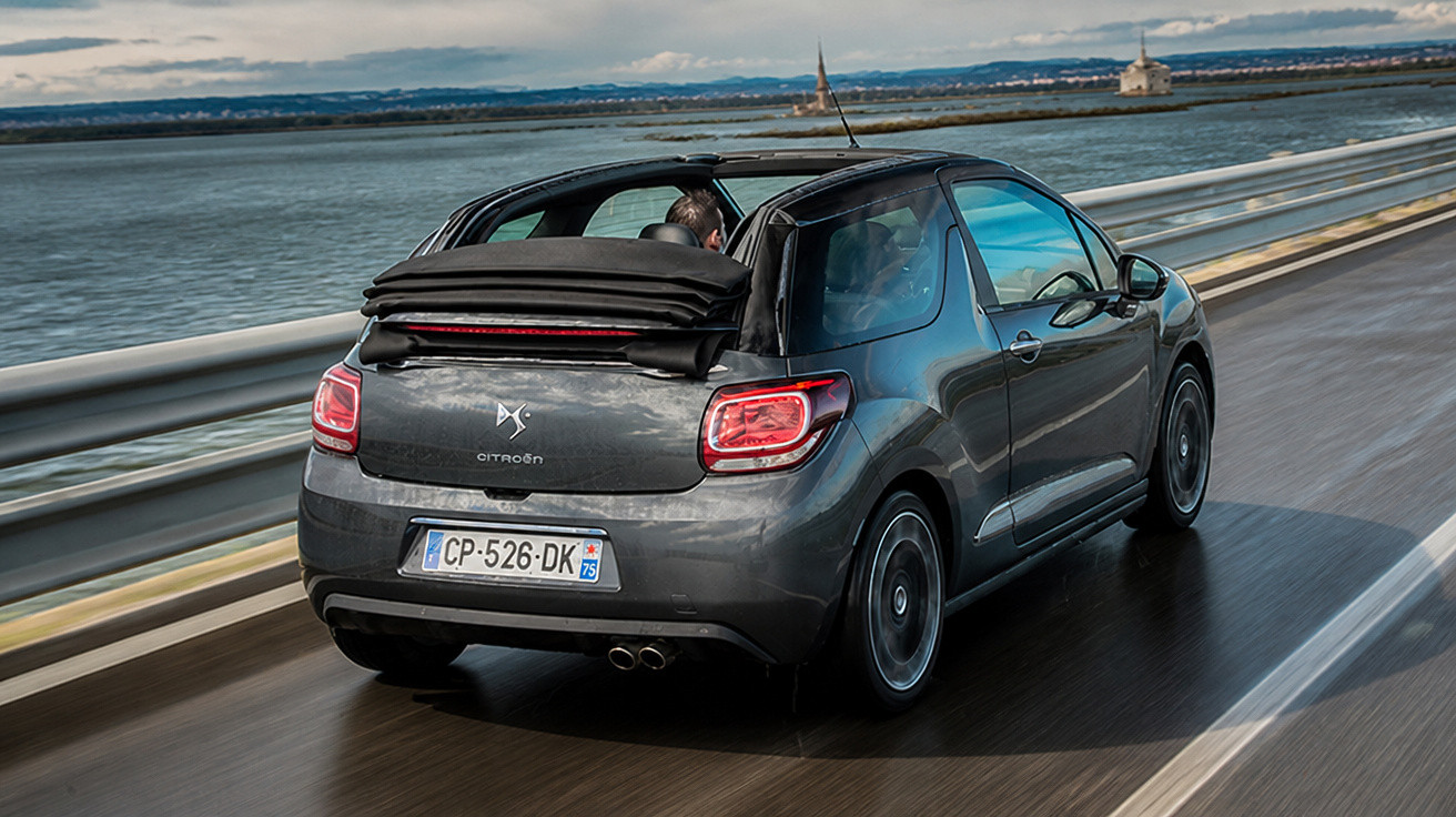 Carro Citroën DS3 Cabrio cinza a conduzir numa estrada à beira-mar com tejadilho aberto.