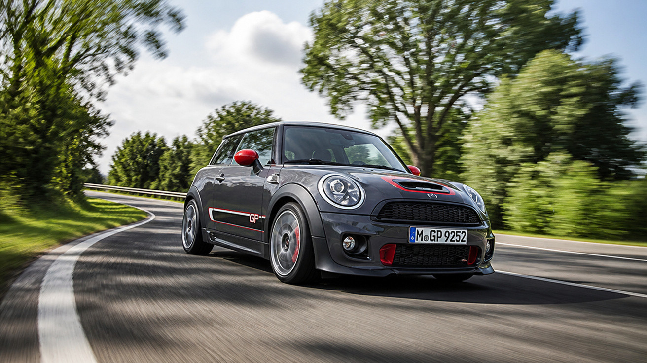 Carro desportivo Mini Cooper cinzento a contornar uma curva numa estrada rodeada de árvores e relva.