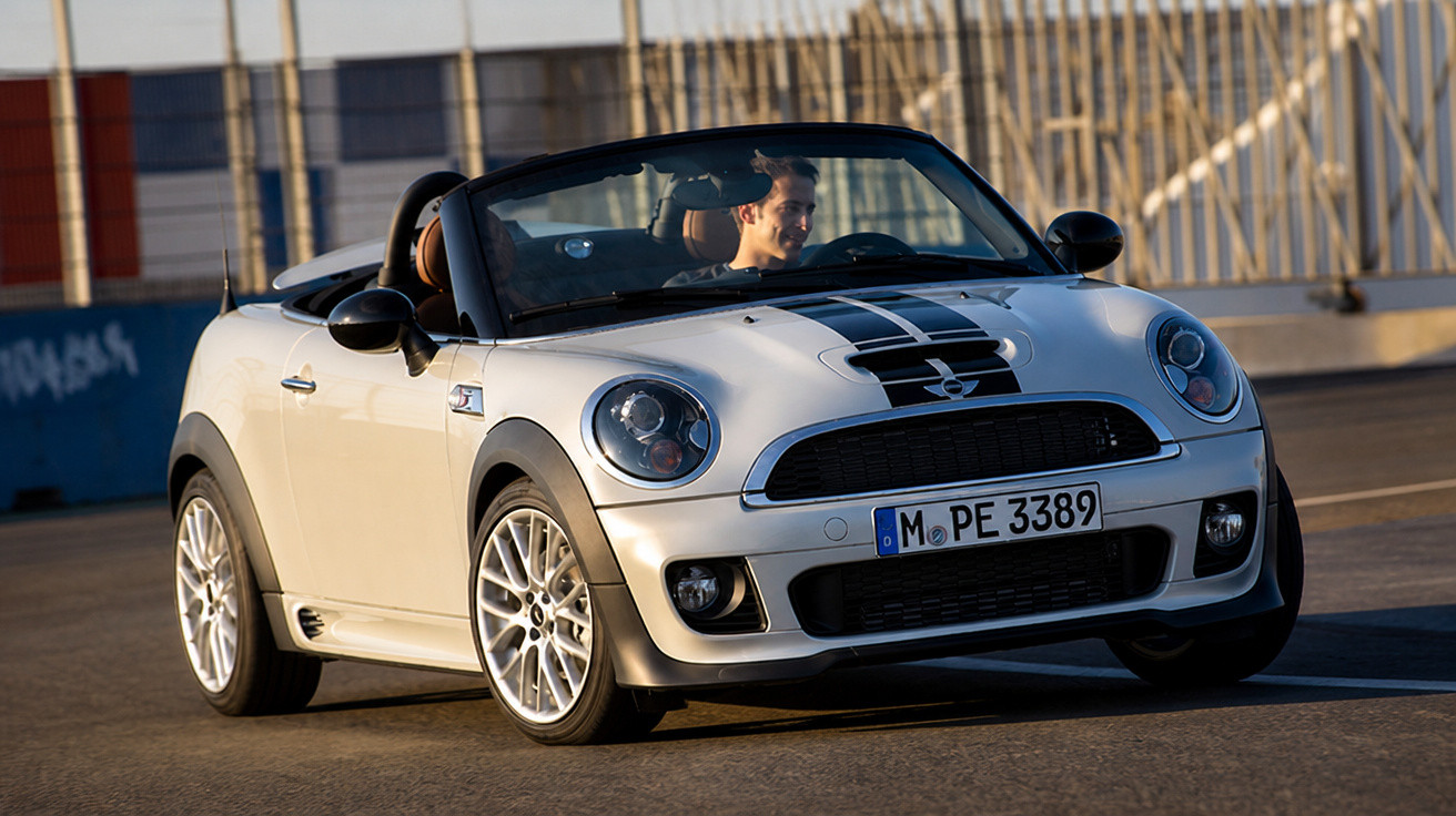 Carro conversível Mini Cooper branco com detalhes pretos a circular numa via urbana com um condutor ao volante.
