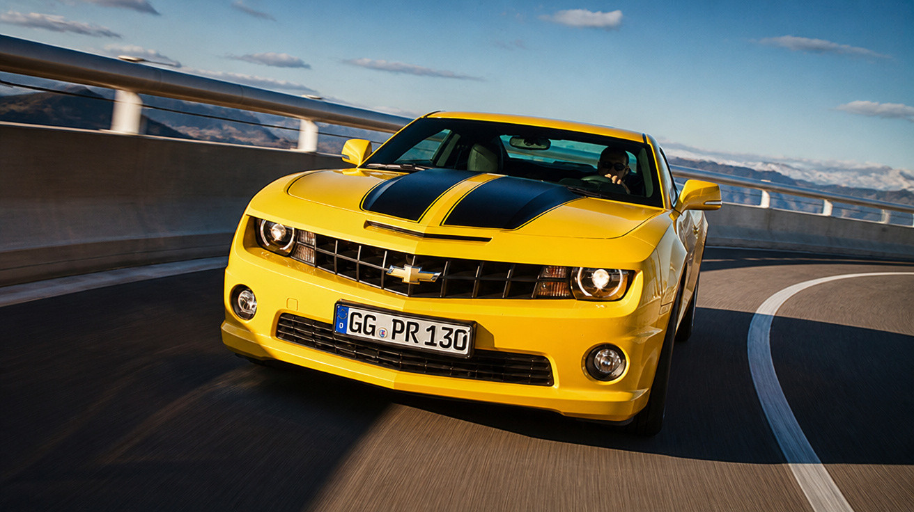 Carro desportivo amarelo com riscas negras a circular numa estrada curvada sob céu azul.