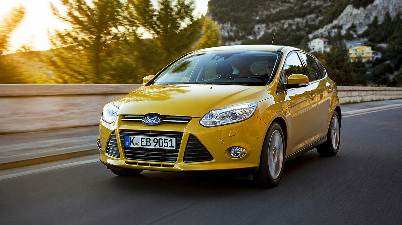 Carro Ford Focus amarelo a circular numa estrada com árvores e montanhas ao fundo ao pôr do sol.