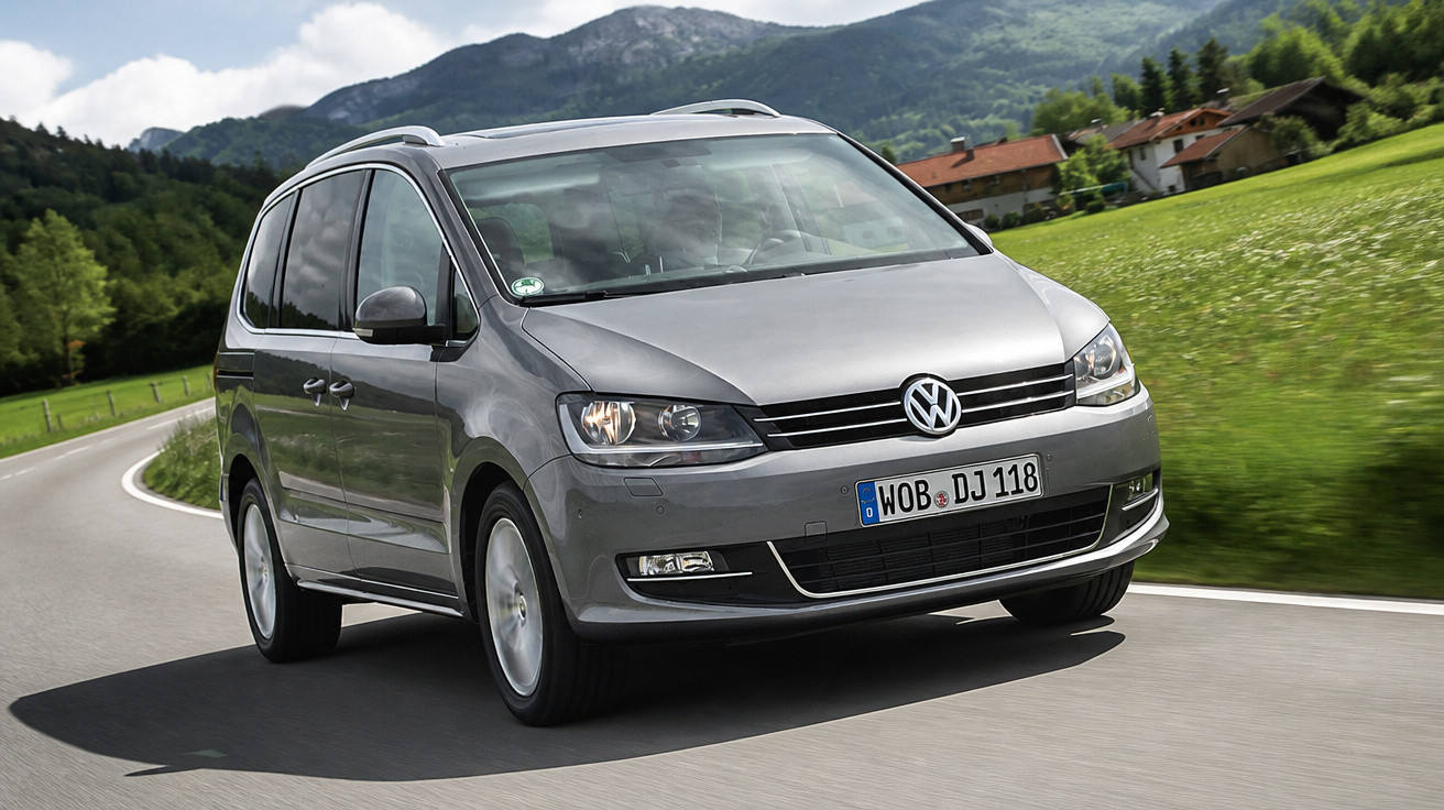 Carro Volkswagen cinzento a circular numa estrada curva rodeada por campo verde e montanhas ao fundo.