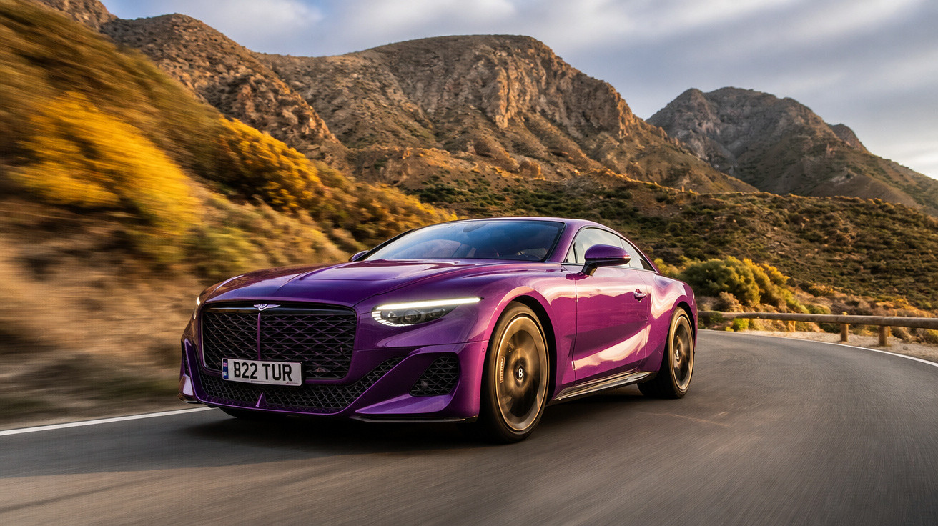 Carro desportivo roxo a circular numa estrada sinuosa entre montanhas ao pôr do sol.