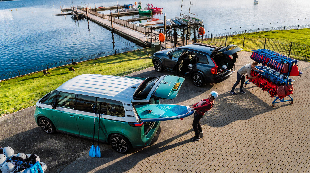 Pessoas a preparar pranchas e coletes junto a carros estacionados perto de um lago com cais e barcos.
