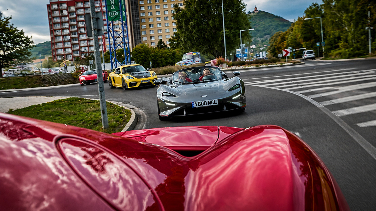 Carros desportivos de luxo a circular em fila numa estrada curva numa zona urbana com edifícios e árvores.
