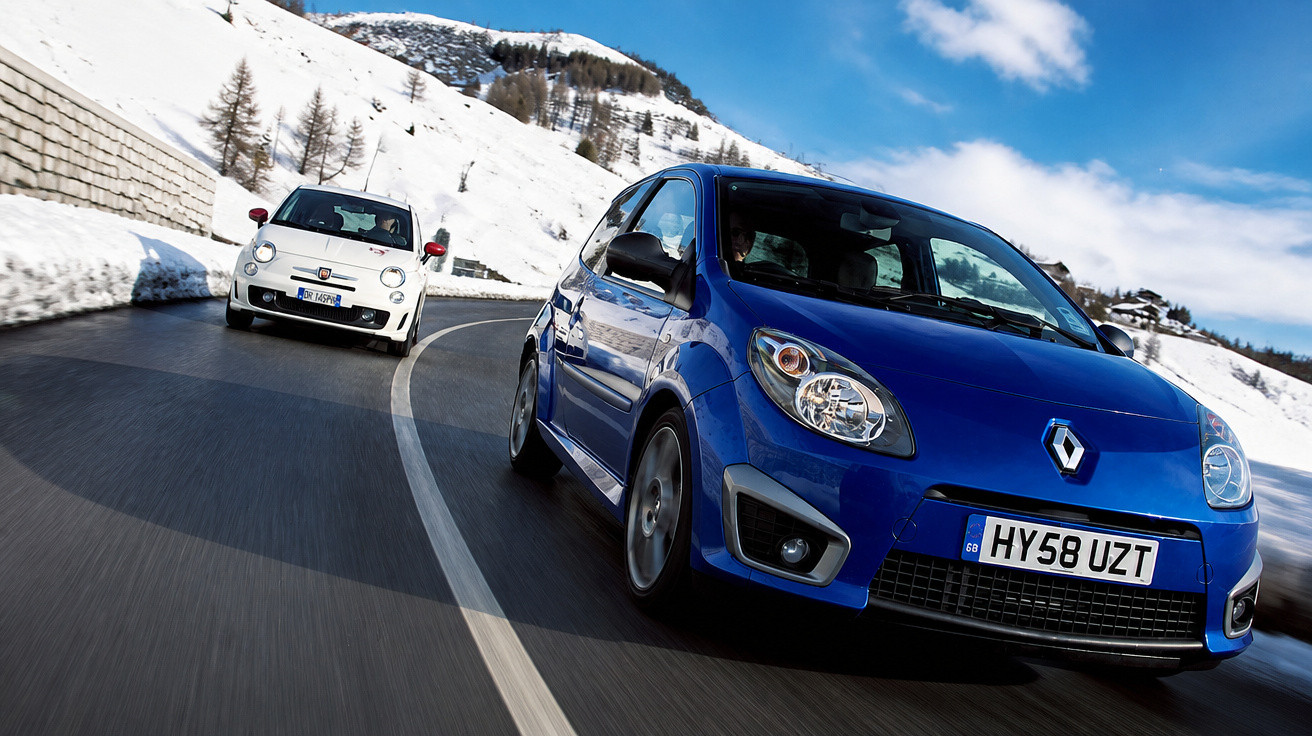 Dois carros urbanos a circular numa estrada montanhosa com neve e céu azul com algumas nuvens.