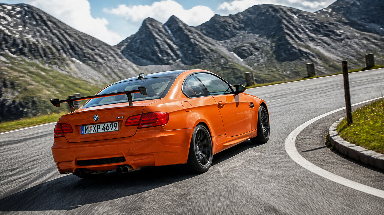 Carro desportivo laranja BMW a acelerar em estrada curva, com montanhas rochosas ao fundo.