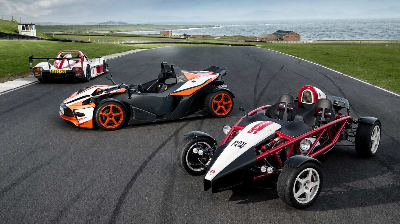 Três carros desportivos estilo buggy num circuito asfaltado junto ao mar, com céu nublado.