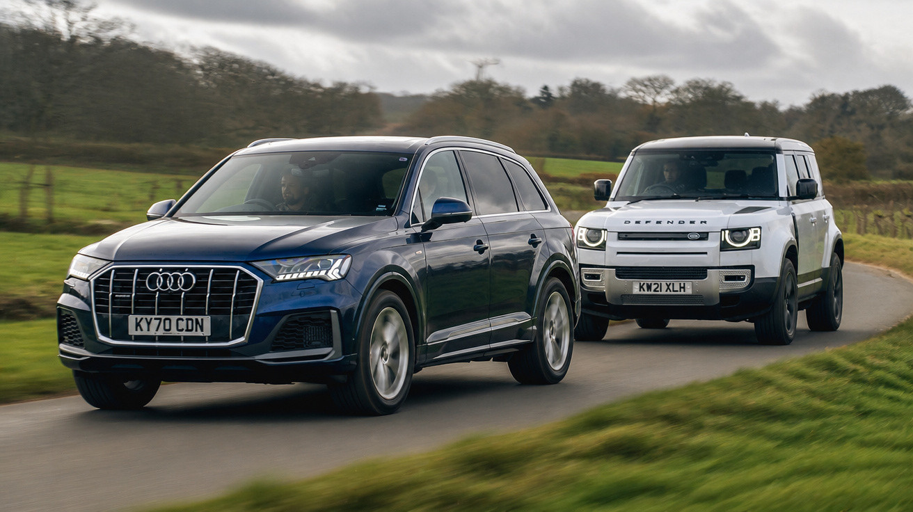Dois SUVs em circulação numa estrada rural, um azul Audi à frente e um branco Land Rover Defender atrás.