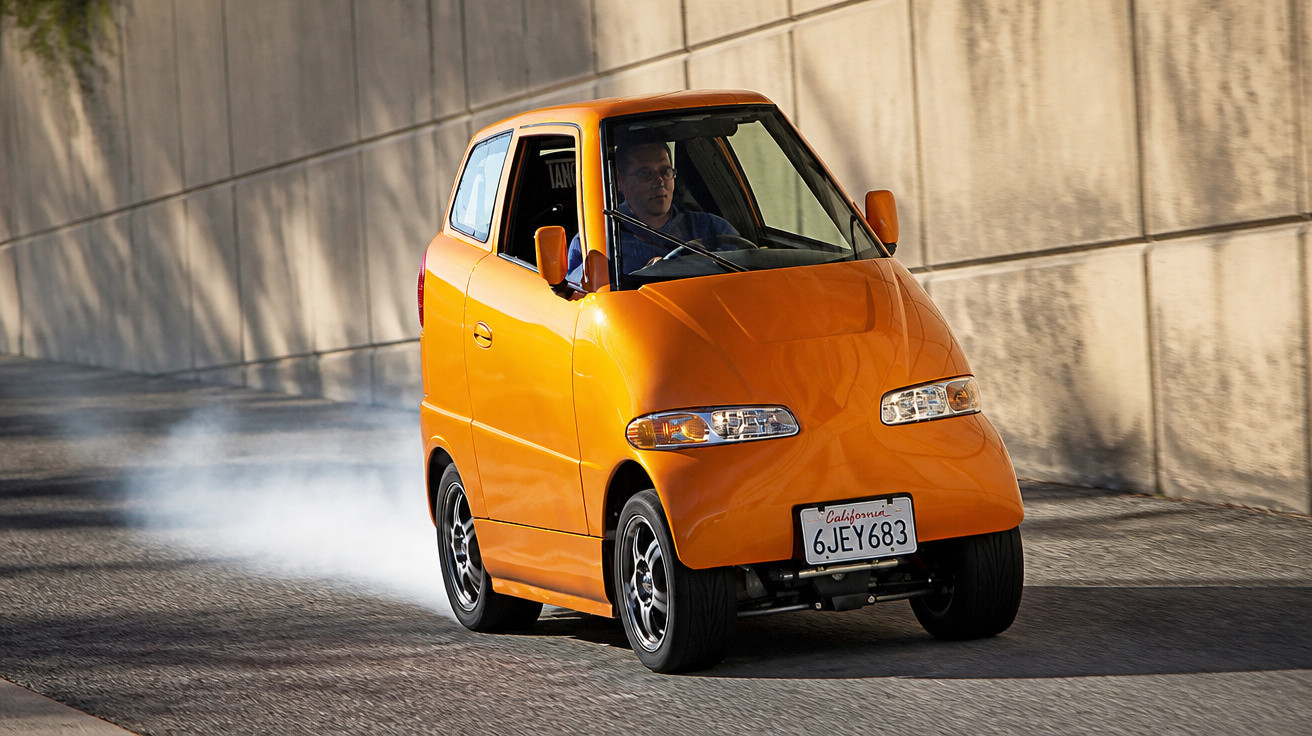 Carro compacto laranja com placa da Califórnia a fazer curva numa estrada com parede de cimento ao lado.