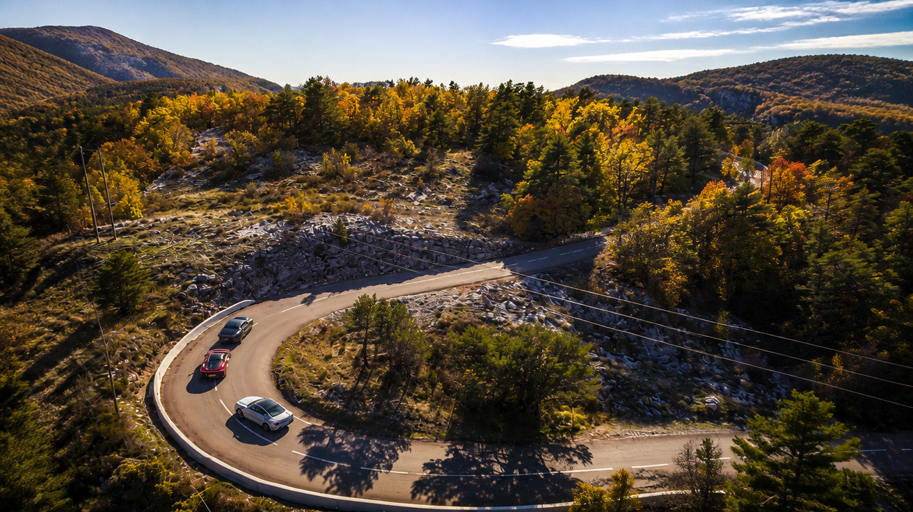 Estrada sinuosa na montanha com três carros a circular rodeada por árvores outonais e céu azul.