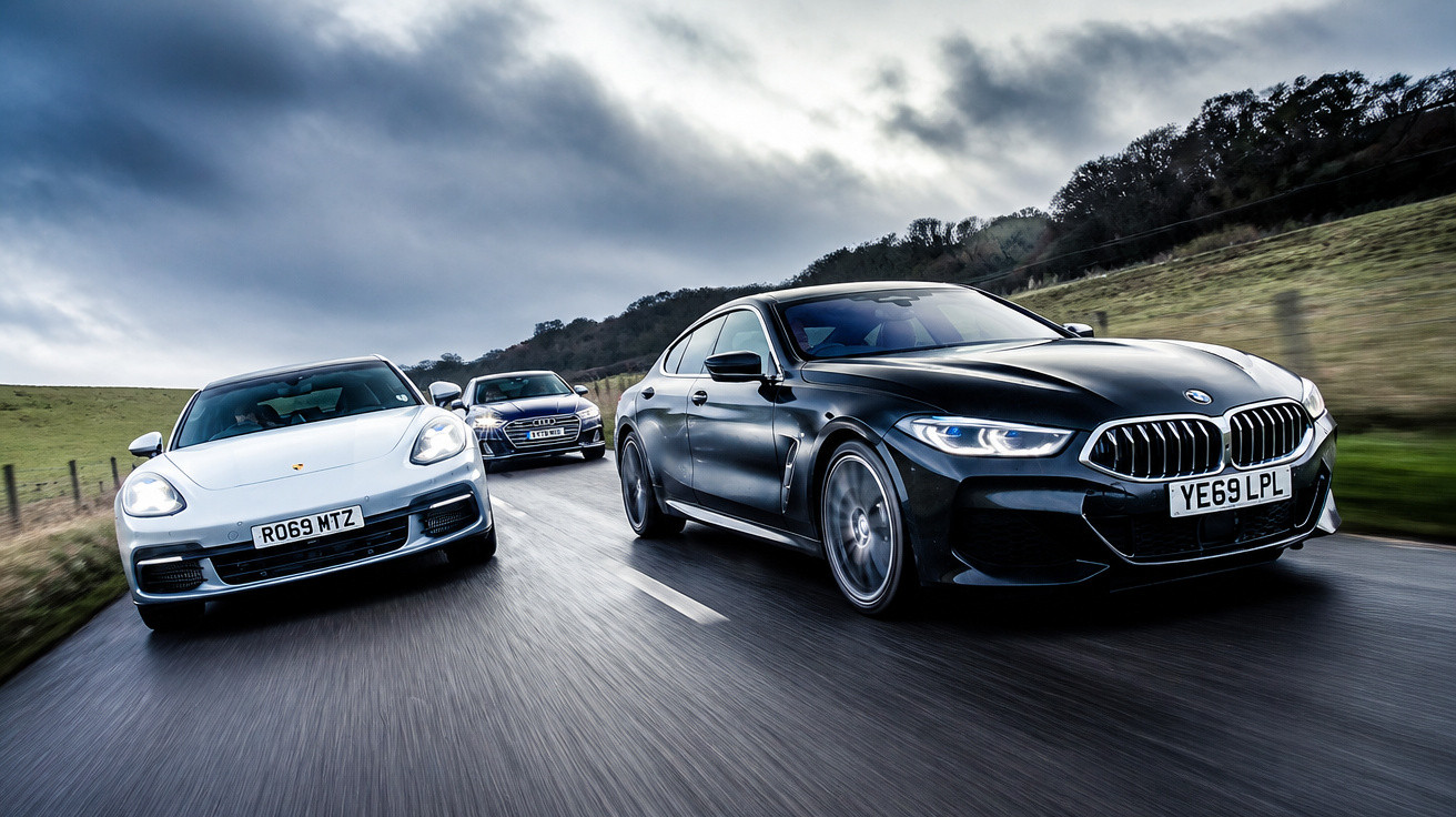 Três carros a alta velocidade numa estrada rural sob céu nublado, com destaque para BMW preto e Porsche branco.