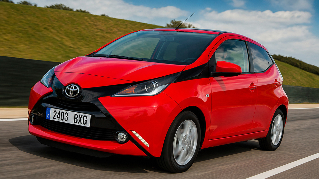 Carro vermelho Toyota Aygo em movimento numa estrada com relva e céu nublado ao fundo.