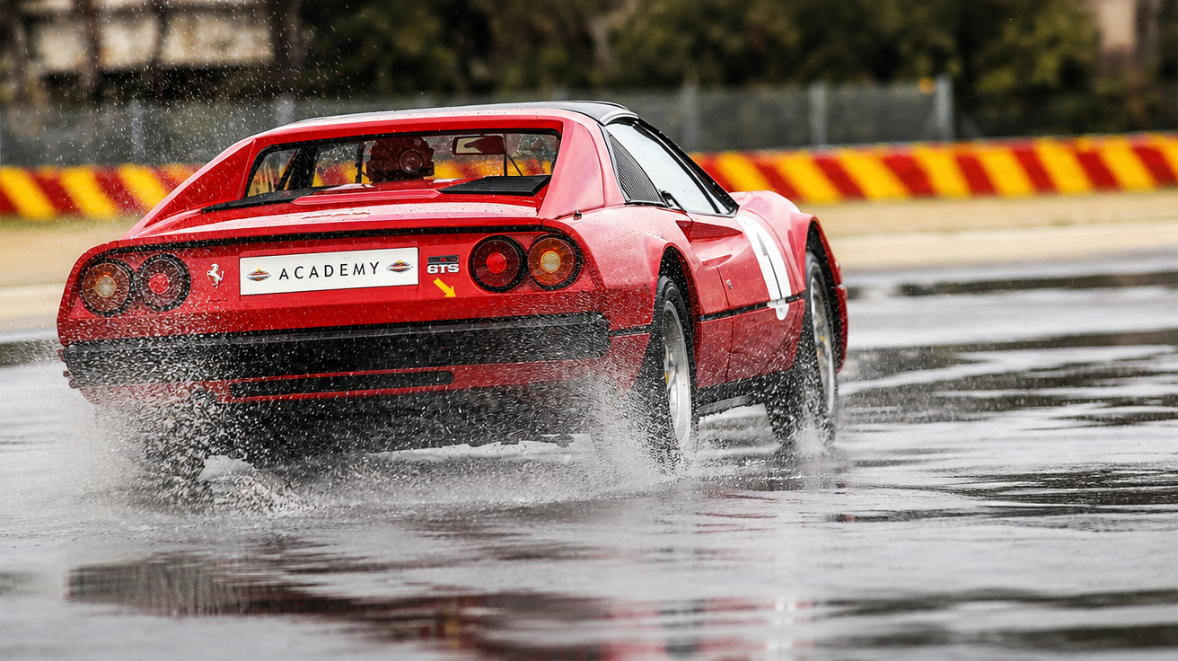 Carro desportivo vermelho a acelerar numa pista molhada, levantando água em volta.